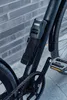 A close-up shot of a black bicycle frame with a black ABUS folding lock attached to it. The background is a brick wall. Náhled obrázku produktu