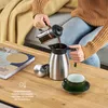 A person is pouring coffee from a French press into a thermos jug, with a cup and saucer nearby. Produktbild-Vorschau