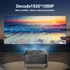 A mini projector is projecting a sunset scene onto a large screen. The projector is black and sits on a table in the foreground. Produktbild-Vorschau
