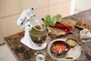 A white KitchenAid stand mixer is shown on a countertop, preparing ingredients for baking. Rhubarb, eggs, flour, and oats are visible, along with a partially prepared rhubarb tart in a baking pan. Produktbild-Vorschau