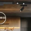 A black spotlight is attached to a track light system on a wooden ceiling. There are also decorative elements and cabinets visible in the background. Produktbild-Vorschau