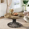 A round wooden coffee table with a metal base is placed on a rug in a living room setting, with a bouquet of flowers and fruit on top. Voir les images du produit