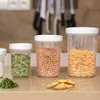 Three clear plastic food storage containers with white lids are arranged on a countertop. The containers hold dried herbs, chickpeas, and pasta. A small pile of dried herbs and some crackers are in front of the containers. Produktbild-Vorschau