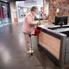 An elderly woman is standing at a checkout counter in a store, holding a shopping basket and a cane. Produktbild-Vorschau