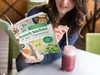 A woman is sitting at a table, holding an open cookbook and looking at it. Next to her is a glass jar with a pink smoothie and a straw. Produktbild-Vorschau