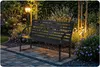 A dark metal garden bench sits on a paved patio in a garden at dusk, illuminated by garden lights. Produktbild-Vorschau