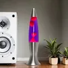 A lava lamp with purple liquid and red lava blobs is placed next to a white speaker and two potted plants. Produktbild-Vorschau