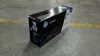 A black HP toner cartridge box sits on a concrete floor with a yellow line painted on it. Voir les images du produit