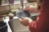 A person is washing a round black item with holes under a faucet in a kitchen sink. Anteprima dell’immagine prodotto