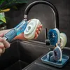 A person is holding a dish soap dispenser and a sponge over a kitchen sink, with a brush holder in the background. Anteprima dell’immagine prodotto