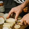 A person is cutting cheese with a knife on a wooden board, with crackers, an apple, and cans of drink in the background. Náhled obrázku produktu