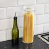A green bottle with a pour spout and a glass jar filled with spaghetti are on a dark countertop in front of a white subway tile backsplash. Produktbild-Vorschau