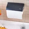 A black paper towel holder mounted on a tiled wall, with a roll of white paper towels dispensing from it. The holder has a small logo on its front. Below the holder, a white countertop is visible, with a decorative glass jar and a portion of some food item Produktbild-Vorschau