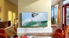 A modern living room with a large TV displaying a surfing image, two orange chairs, and a window with curtains. Produktbild-Vorschau