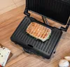 A panini grill with a sandwich on it, placed on a wooden table. Produktbild-Vorschau