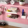 A pink pegboard desk organizer with various items like headphones, keys, envelopes, a glasses case, pens, and a camera. Voir les images du produit