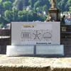 A product box and filters are placed on a stone ledge with a blurred background of trees and buildings. Produktbild-Vorschau