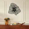 A decorative metal wall clock shaped like a flower is mounted on a wall above a wooden console table. The table holds a vase of flowers and some decorative items. Produktbild-Vorschau