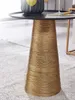 A close-up shot of a round side table with a gold, textured base and a black tabletop, placed on a patterned rug. Náhled obrázku produktu