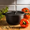 A cast iron pot with a lid is placed on a wooden table, surrounded by red bell peppers, bay leaves, and peppercorns. The background features a tiled wall. Náhled obrázku produktu