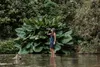 A woman stands in shallow water, drinking from a water bottle, with lush green foliage behind her. Podgląd zdjęcia produktu
