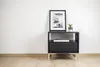 A black nightstand with a framed picture and a small plant on top, placed against a white wall and wooden floor. Produktbild-Vorschau