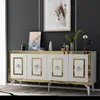 A white and gold sideboard with decorative elements is displayed against a dark gray wall, with artwork and decorative items on top. Produktbild-Vorschau