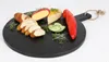A round black wooden serving board with various food items arranged on it, including sliced bread, cheese, cucumber slices, sprouts, and a red pepper. Anteprima dell’immagine prodotto