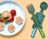 A plate of food shaped like a caterpillar, next to a set of children's cutlery. Produktbild-Vorschau