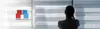 A person is looking out a window with blinds. A logo is present on the left side. Produktbild-Vorschau