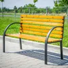 A wooden park bench with black metal legs sits on a paved walkway, with grass and a fence in the background. Anteprima dell’immagine prodotto