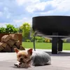 A small dog is lying on a wooden deck next to a pile of firewood and a large black fire pit. Produktbild-Vorschau