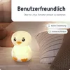 A cute, glowing penguin-shaped night light is placed on a bed with pillows. Produktbild-Vorschau