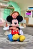 A young girl is sitting on the floor, hugging a large Mickey Mouse plush toy in a bedroom. Voir les images du produit