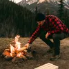 A man is adding wood to a campfire in a forest with mountains in the background. Produktbild-Vorschau