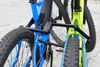 Close-up view of the rear wheels and frame of two bicycles, one blue and one green, connected by a black folding lock mechanism. Produktbild-Vorschau