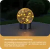 A solar-powered decorative light with LED lights sits on a table outdoors at night. Produktbild-Vorschau
