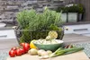 A kitchen counter with fresh ingredients including rosemary, tomatoes, potatoes, lemons, and green onions, arranged around a basket of rosemary. Produktbild-Vorschau