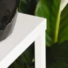 A close-up view of a white plant stand with a black pot and green leaves in the background. Anteprima dell’immagine prodotto