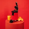 A red electric juicer is displayed on a red pedestal with a glass of orange juice and oranges next to it, against a red background. Podgląd zdjęcia produktu