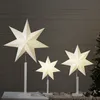 Three illuminated star-shaped lamps of varying sizes are displayed on a table against a wooden plank background. A portion of a Christmas tree is visible on the right. Podgląd zdjęcia produktu