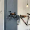 A close-up of a bicycle-shaped pizza cutter mounted on a gray column, with another bicycle frame visible in the background. Produktbild-Vorschau