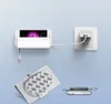 A white smartphone charging holder is mounted on a light blue wall, with a smartphone plugged in and charging. A power adapter is plugged into a wall socket. Náhled obrázku produktu