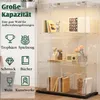 A clear acrylic display cabinet with three shelves, showcasing various items like books, decorative objects, and a vintage radio. The cabinet has integrated LED lighting. Produktbild-Vorschau