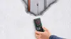 A hand holding a Bosch Truvo digital detector against a textured wall with a hole revealing pipes and wires. Voir les images du produit