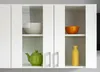 A close-up view of a white kitchen cabinet with two shelves. The top shelf holds a light-colored bowl, and the bottom shelf holds a green teapot. The right side of the cabinet has an orange bowl on the top shelf and a yellow vase on the bottom shelf. Náhľad obrázku produktu
