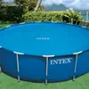 An outdoor shot of a round, blue Intex swimming pool covered with a blue solar cover. The pool is situated on a green lawn, with lush green bushes and palm trees in the background. Mountains and a body of water are visible in the distance under a cloudy sk Voir les images du produit