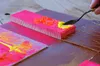A close-up shot of someone using a palette knife to apply yellow acrylic paint to a pink wooden block, with other painted surfaces visible in the background. Podgląd zdjęcia produktu