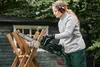 A woman is using an electric chainsaw to cut a log in an outdoor setting. Produktbild-Vorschau