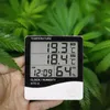 A hand holds a digital thermometer and hygrometer in front of lush green foliage. Anteprima dell’immagine prodotto